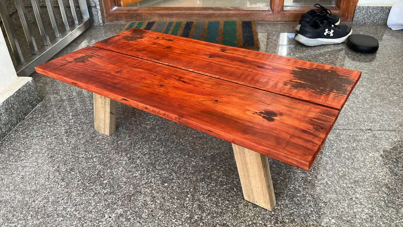 A low hand-built bench with a scorched, red-orange plank top on pale wooden legs.