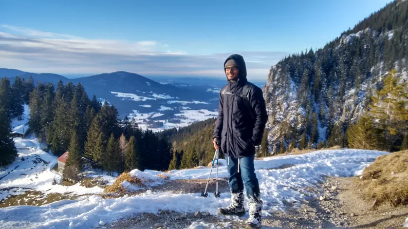 Jason standing on a snowy mountain trail with pine forest and valley views behind.
