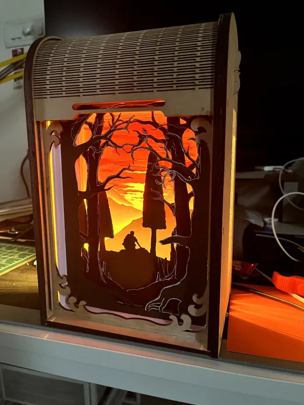 A backlit, laser-cut wooden diorama box showing a silhouetted figure among trees at sunset.